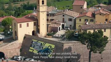 Widok z lotu ptaka na włoską wioskę Tavullia, z widocznym napisem "WELCOME TO TAVULLIA" na ścianie budynku.