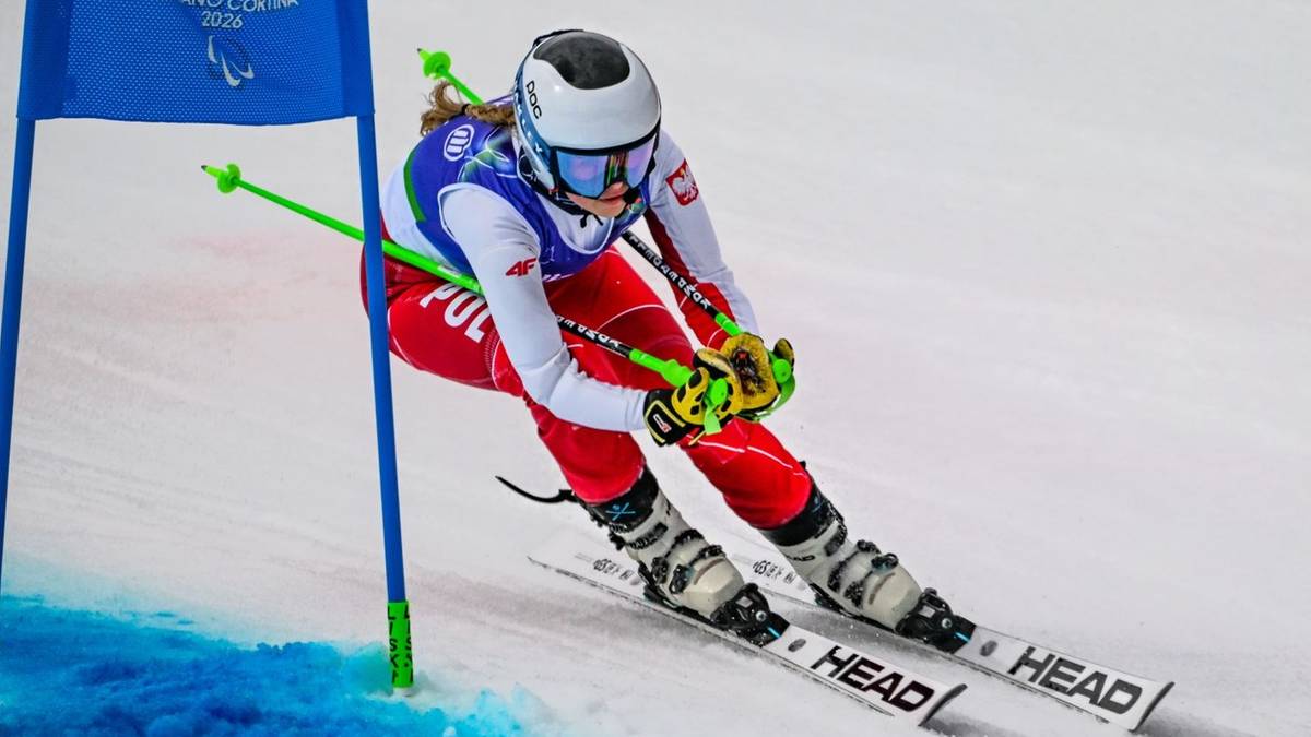 Narciarka alpejska w czerwonym stroju i białym kasku z zielonymi kijkami zjeżdża po stoku narciarskim.