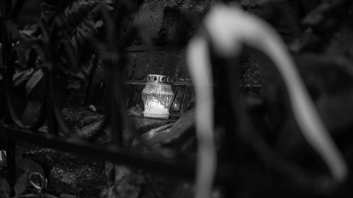 Szklany znicz zapalony wśr&oacute;d kamieni i ciemnych roślin, z rozmytą postacią w tle.