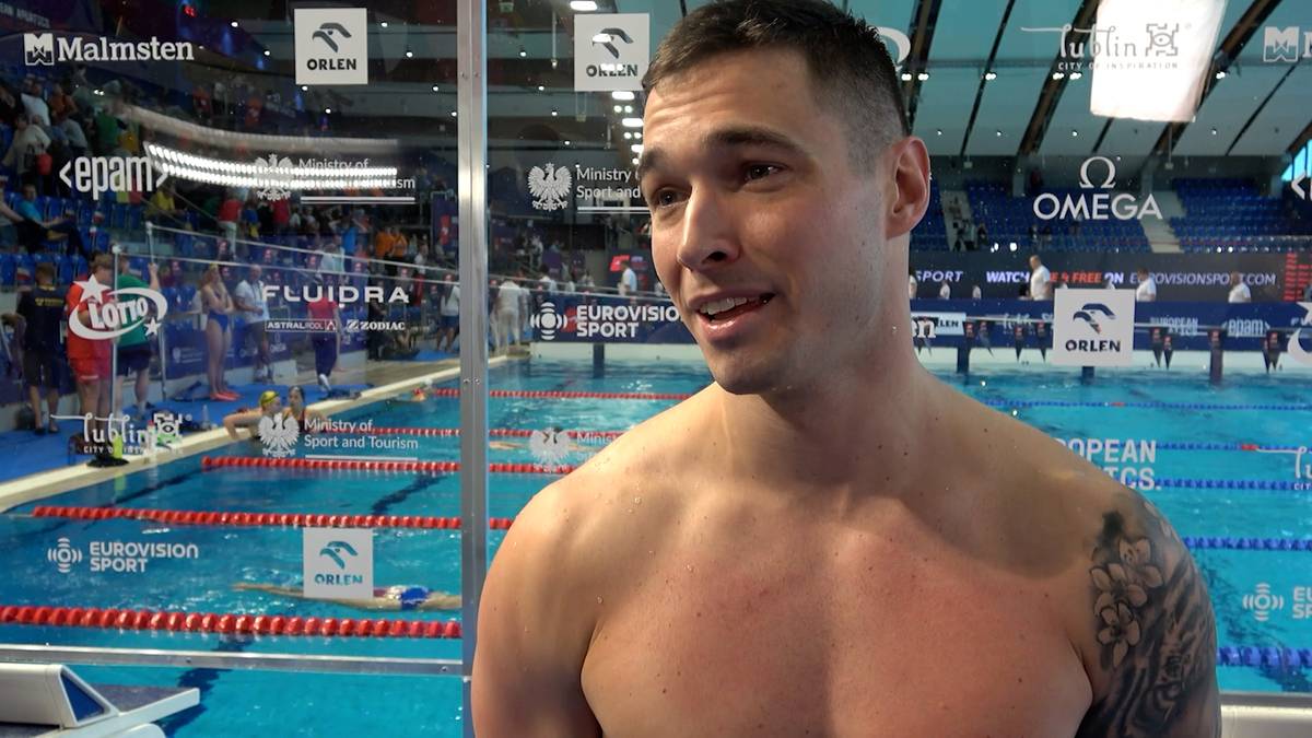 Uśmiechnięty mężczyzna, z widocznym tatuażem na ramieniu, stoi na tle basenu pływackiego.