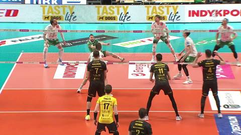 Siatkarz w białej koszulce z numerem i logo, z pochyloną głową, patrzy w d&oacute;ł. W tle widzowie na trybunach i inni gracze.