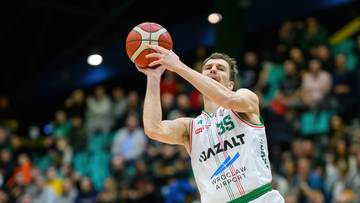 Koszykarz w białym stroju z numerem 35 rzuca do kosza, broniony przez zawodnika w niebieskim stroju.