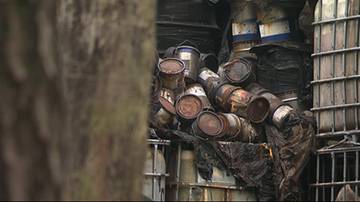 Stare, zardzewiałe beczki i pojemniki stojące w nieporządku, częściowo przykryte czarną folią.