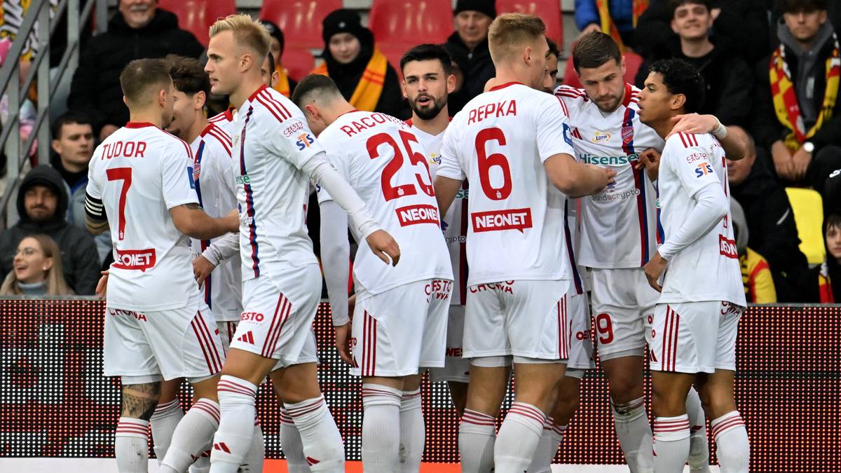 Grupa piłkarzy Rakowa Częstochowa w białych strojach z czerwonymi detalami, stojących w kole na boisku.