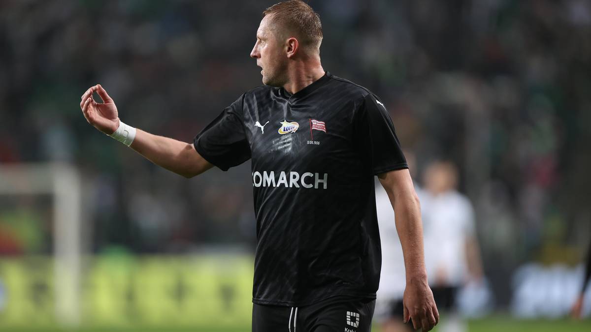 Piłkarz w czarnej koszulce z napisem "OMARCH" i białą opaską na nadgarstku, stojący na boisku.