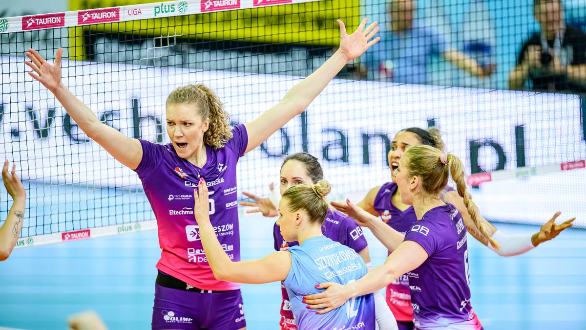 Pięć siatkarek w fioletowych strojach świętuje na boisku siatkarskim, z siatką w tle.