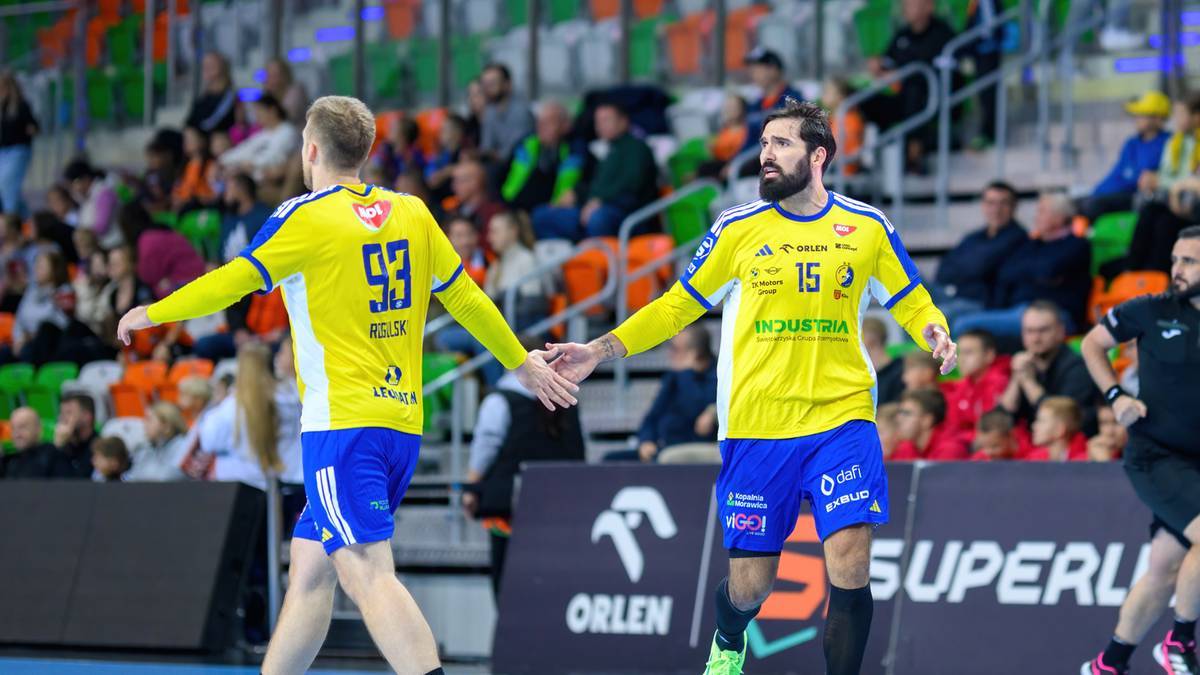 Dwóch zawodników Industrii Kielce w żółtych koszulkach z numerami 93 i 15, wychodzi na boisko podczas meczu Ligi Mistrzów. Jeden z zawodników wykonuje gest dłonią. Na trybunach widoczni są kibice.