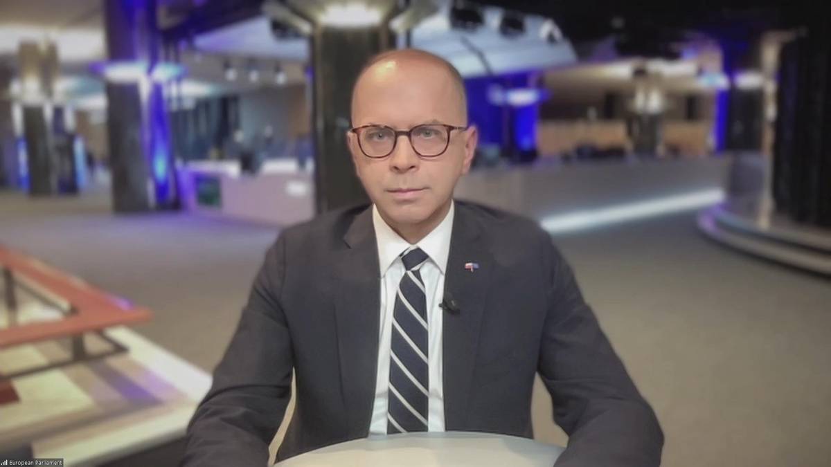 Mężczyzna w okularach i garniturze siedzi przed rozmytym tłem studia telewizyjnego.