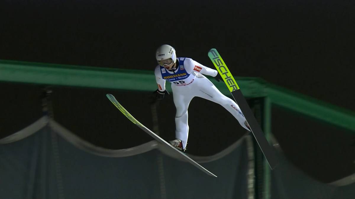 Skoczek narciarski w locie, w białym kombinezonie i kasku, na tle ciemnego nocnego nieba i konstrukcji skoczni.