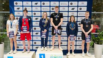 Pięcioro młodych pływak&oacute;w stoi na podium w strojach sportowych, trzymając medale i dyplomy. Są oni zwycięzcami zawod&oacute;w pływackich.