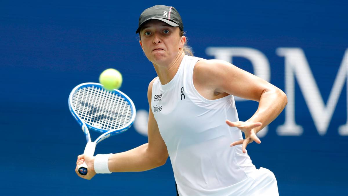 Tenisistka Iga Świątek w akcji podczas meczu, w białej koszulce i czapce, odbijająca piłkę rakietą.