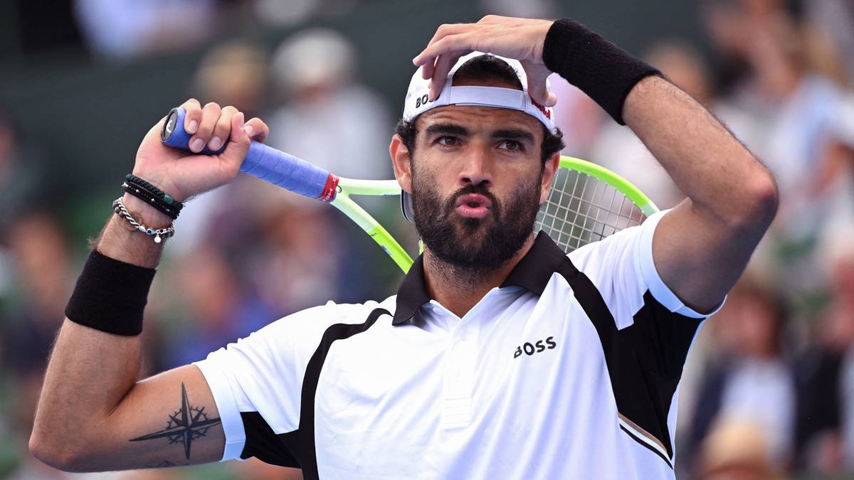 Tenisista Matteo Berrettini w białej koszulce z czarnymi wstawkami i czapką z daszkiem, trzymający rakietę tenisową.