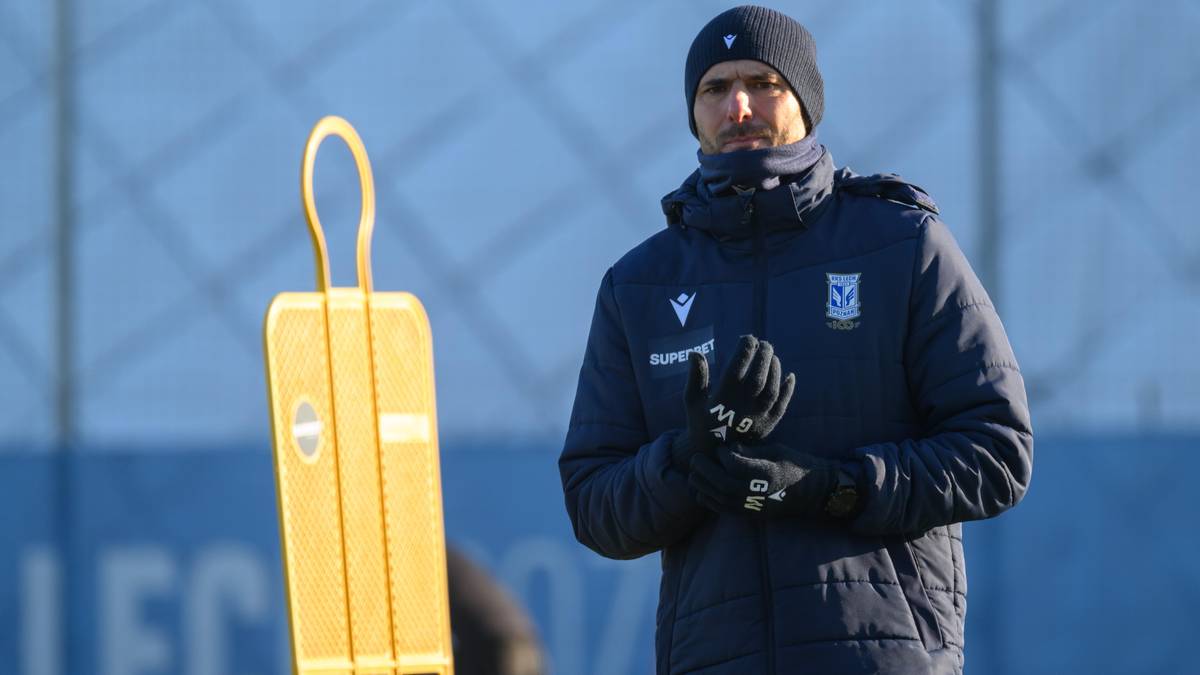 Trener piłkarski w czapce i kurtce sportowej stoi na boisku obok żółtego pachołka treningowego.