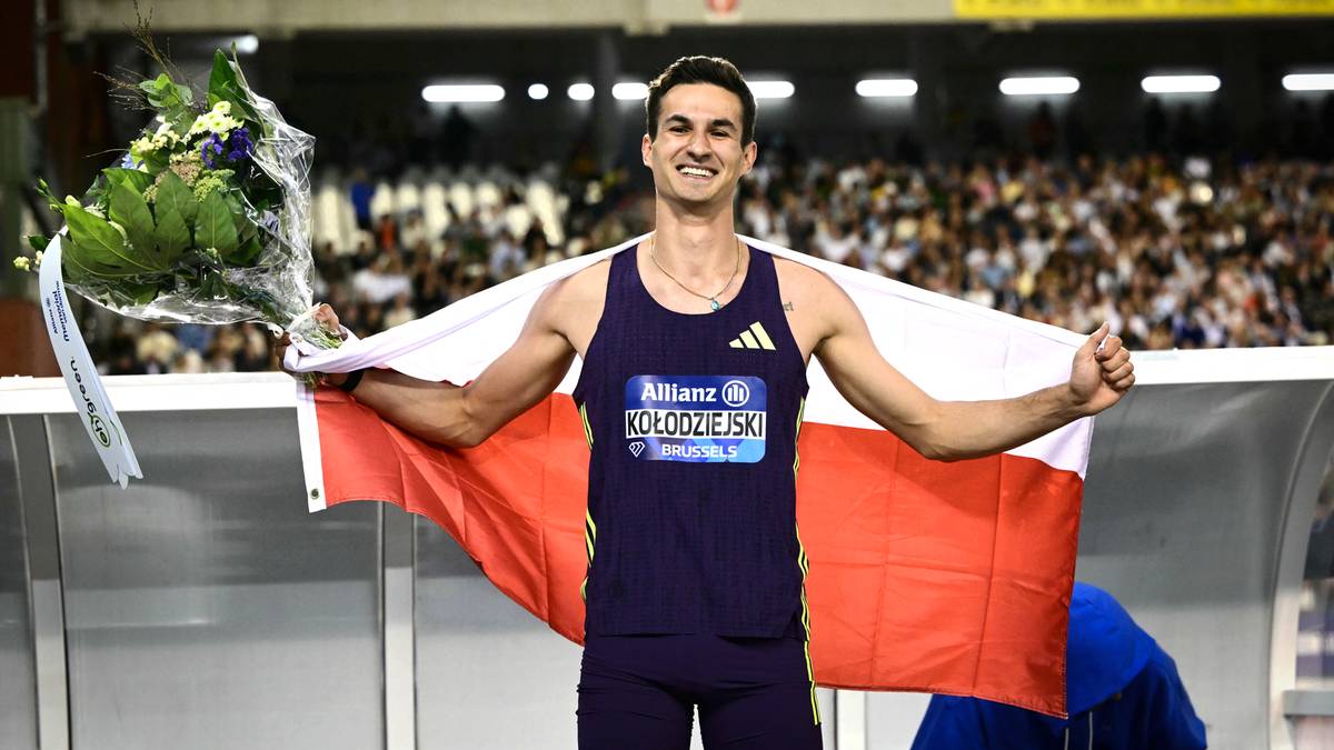 Lekkoatleta w granatowym stroju sportowym z biało-czerwoną flagą zarzuconą na plecy, trzymający bukiet kwiat&oacute;w. Mężczyzna stoi na stadionie, uśmiecha się do obiektywu.