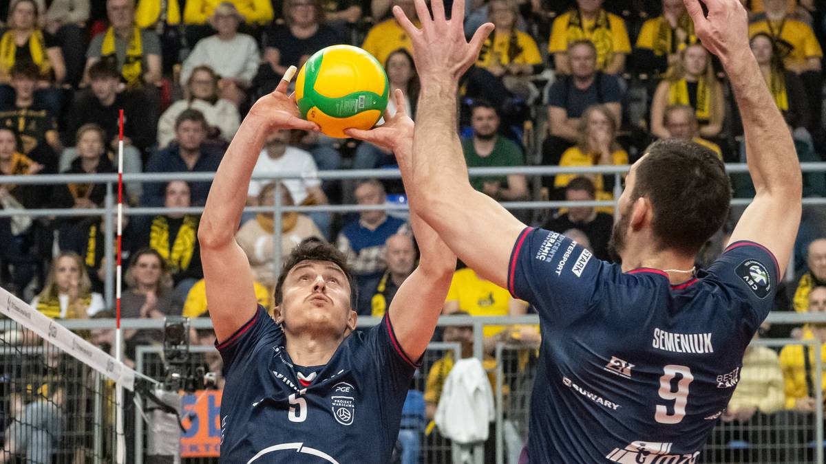 Dw&oacute;ch siatkarzy w ciemnych strojach sportowych walczy o zagranie piłki siatkowej, z publicznością na trybunach w tle.