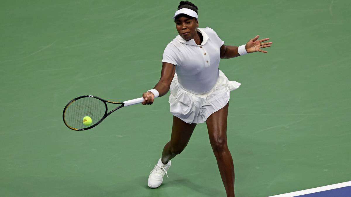 Venus Williams uderza piłkę rakietą tenisową na zielonym korcie, w białej koszulce polo, białej sp&oacute;dniczce i czarnej opasce na głowie.