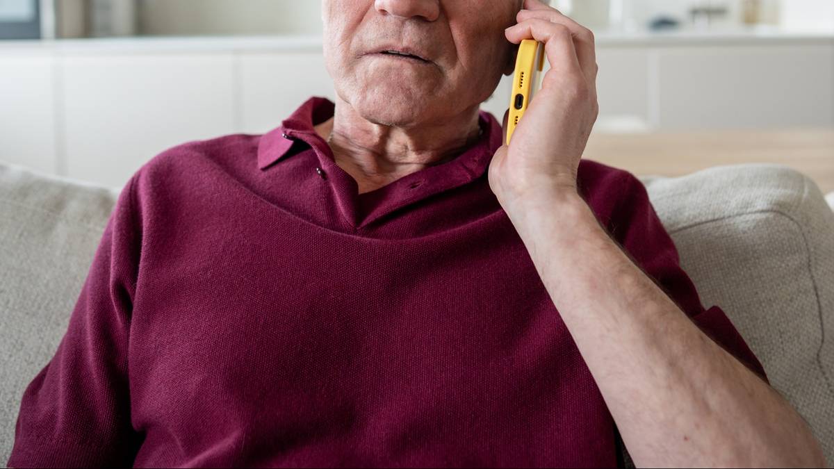 Starszy mężczyzna w bordowej koszuli rozmawia przez ż&oacute;łty telefon kom&oacute;rkowy, siedząc na kanapie.