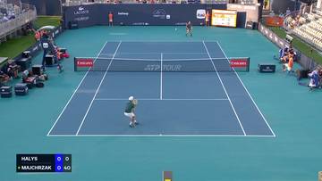 Gracz w zielonej koszulce i białej czapce tenisowej odbija piłkę na niebieskim korcie tenisowym podczas meczu ATP Tour. Wynik na tablicy pokazuje, że Quentin Halys prowadzi 0-0, a Kamil Majchrzak 0-40.