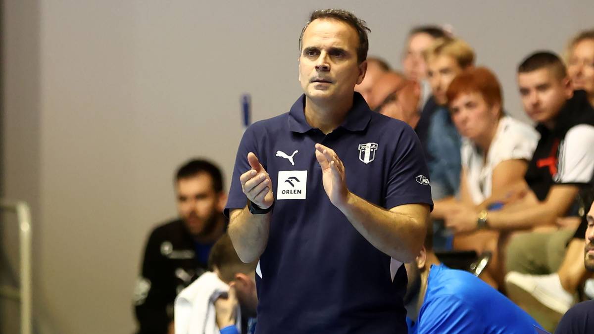 Trener w granatowej koszulce polo z dłońmi złożonymi przed sobą, stojący na tle trybun z kibicami.