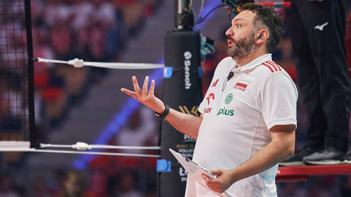 Trener siatk&oacute;wki w białej koszulce z czerwonymi pasami, z białą opaską na ręce, gestykuluje prawą ręką i trzyma kartkę w lewej.