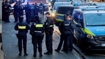 Grupa policjant&oacute;w w ciemnych mundurach z ż&oacute;łtymi elementami odblaskowymi stoi na ulicy obok radiowoz&oacute;w.