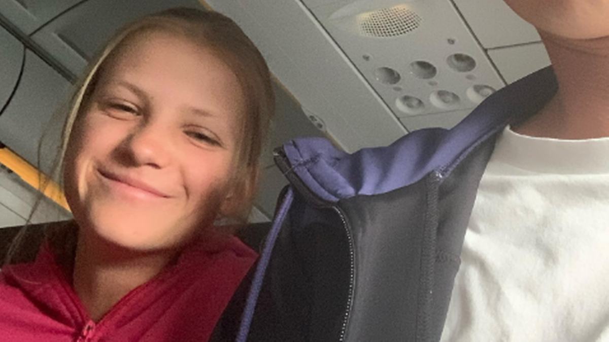 Młoda kobieta uśmiecha się do kamery siedząc w samolocie obok drugiej osoby, której plecy są widoczne.