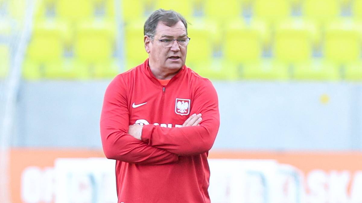 Jan Urban, trener w czerwonej bluzie z herbem reprezentacji Polski, stoi z rękami skrzyżowanymi na piersi.