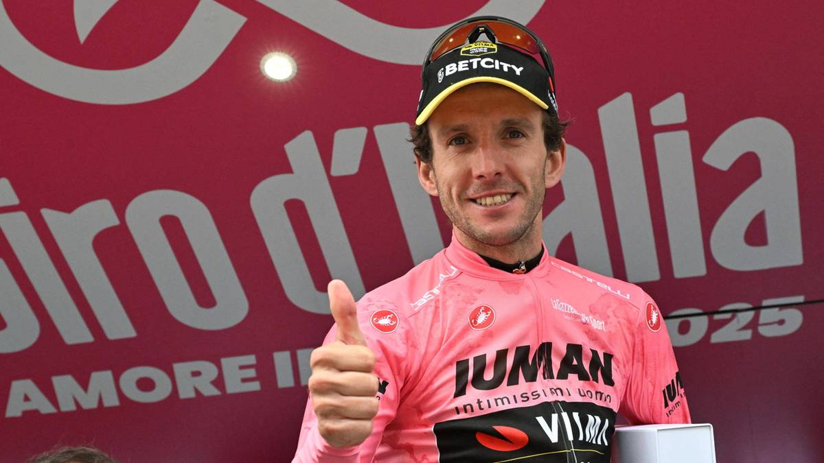 Uśmiechnięty mężczyzna w różowej koszulce kolarskiej z napisem JUMAN, z kciukiem w górę, stoi przed różowym tłem z napisem Giro d'Italia.