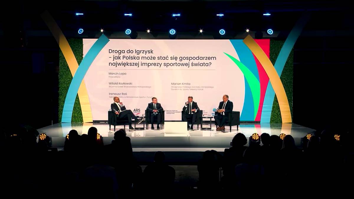 Czterech mężczyzn w garniturach siedzi na krzesłach na scenie, podczas panelu dyskusyjnego na tle ekranu z napisem "Droga do igrzysk - czy Polska może stać się gospodarzem największej imprezy sportowej świata?". Scena jest oświetlona.