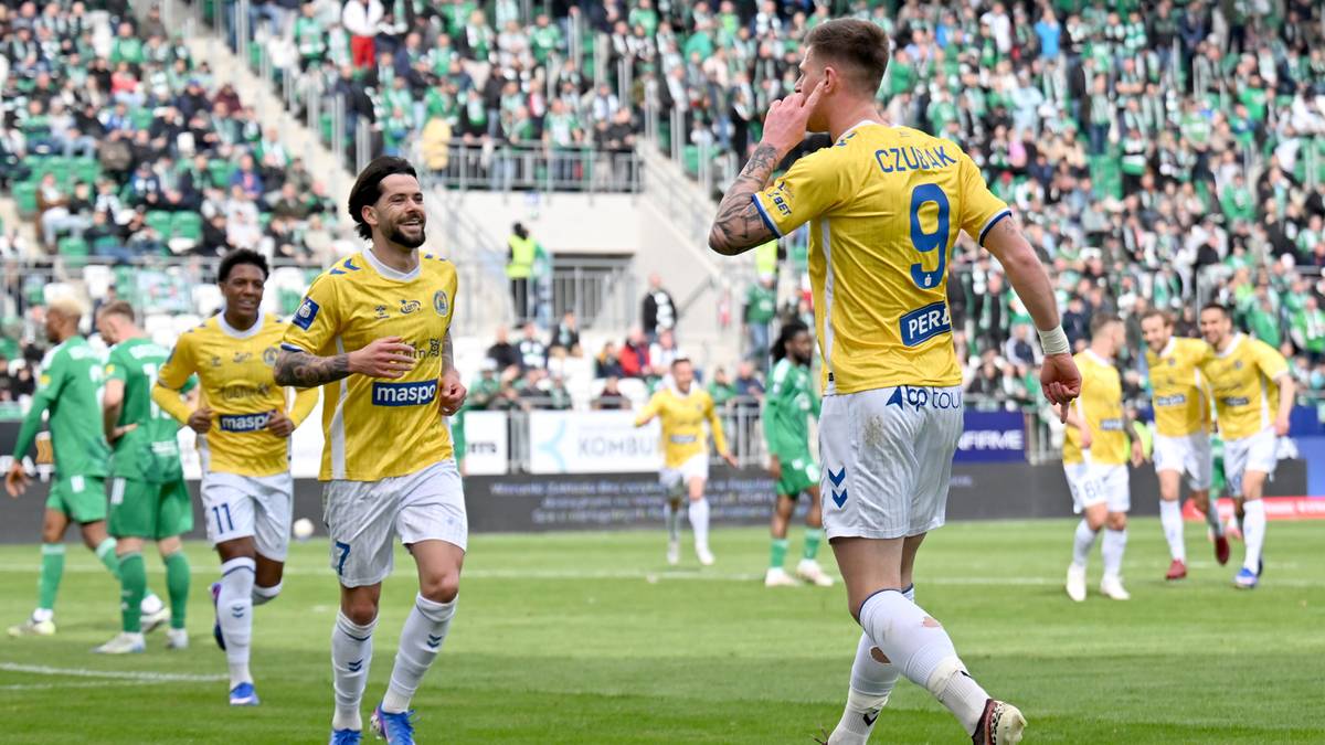 Dwóch piłkarzy w żółtych strojach świętuje na boisku piłkarskim, jeden z nich ma rękę przy uchu.