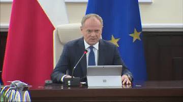 Donald Tusk siedzi za stołem i m&oacute;wi do mikrofonu. Za nim wiszą flagi Polski i Unii Europejskiej.