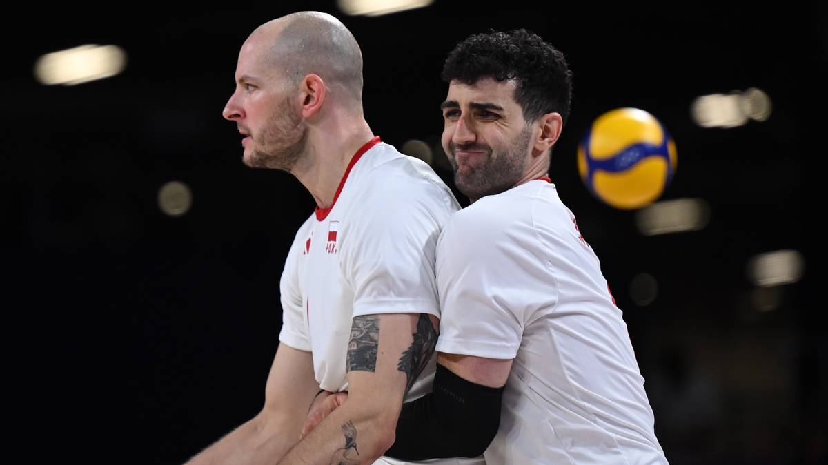 Dwóch siatkarzy w białych koszulkach, jeden z nich podtrzymuje drugiego od tyłu. W tle widoczna jest piłka siatkowa.