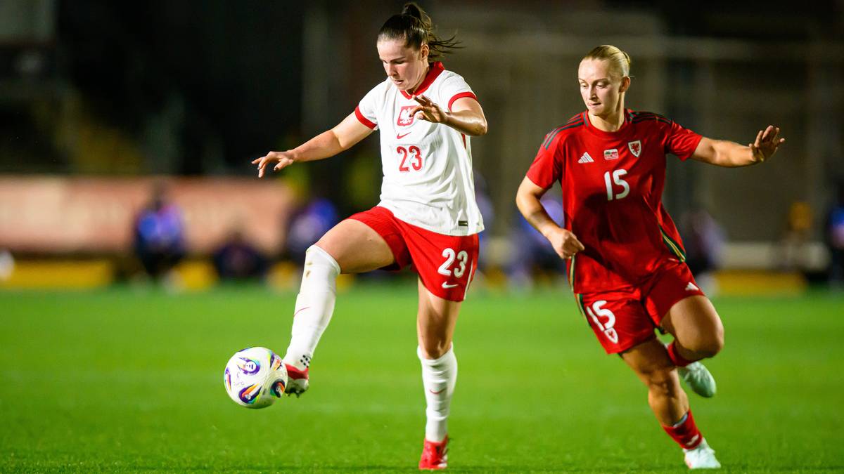 Piłkarka Polski w białej koszulce z numerem 23 i czerwonymi spodenkami prowadzi piłkę, a zawodniczka przeciwniej drużyny w czerwonej koszulce z numerem 15 próbuje ją zatrzymać.