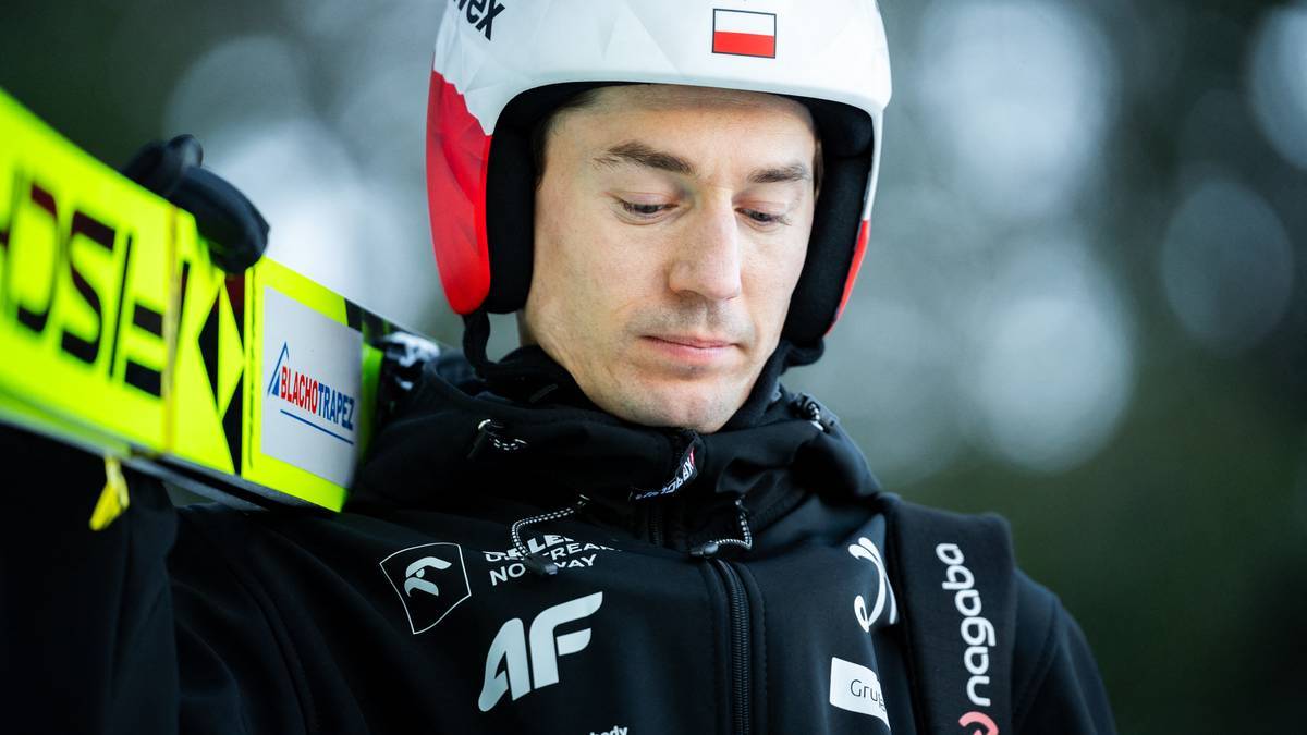 Skoczek narciarski Kamil Stoch w białym kasku z polską flagą, trzymający narty na ramieniu.