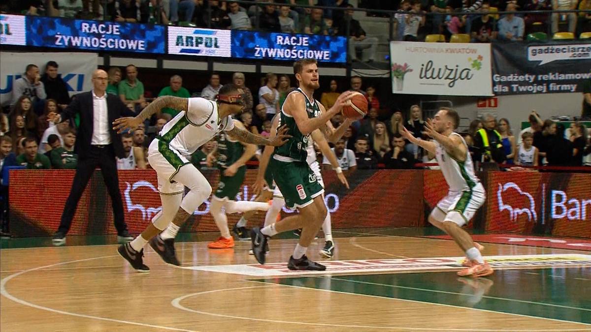 Koszykarz w zielonym stroju wykonuje rzut do kosza, otoczony przez dwóch przeciwników.