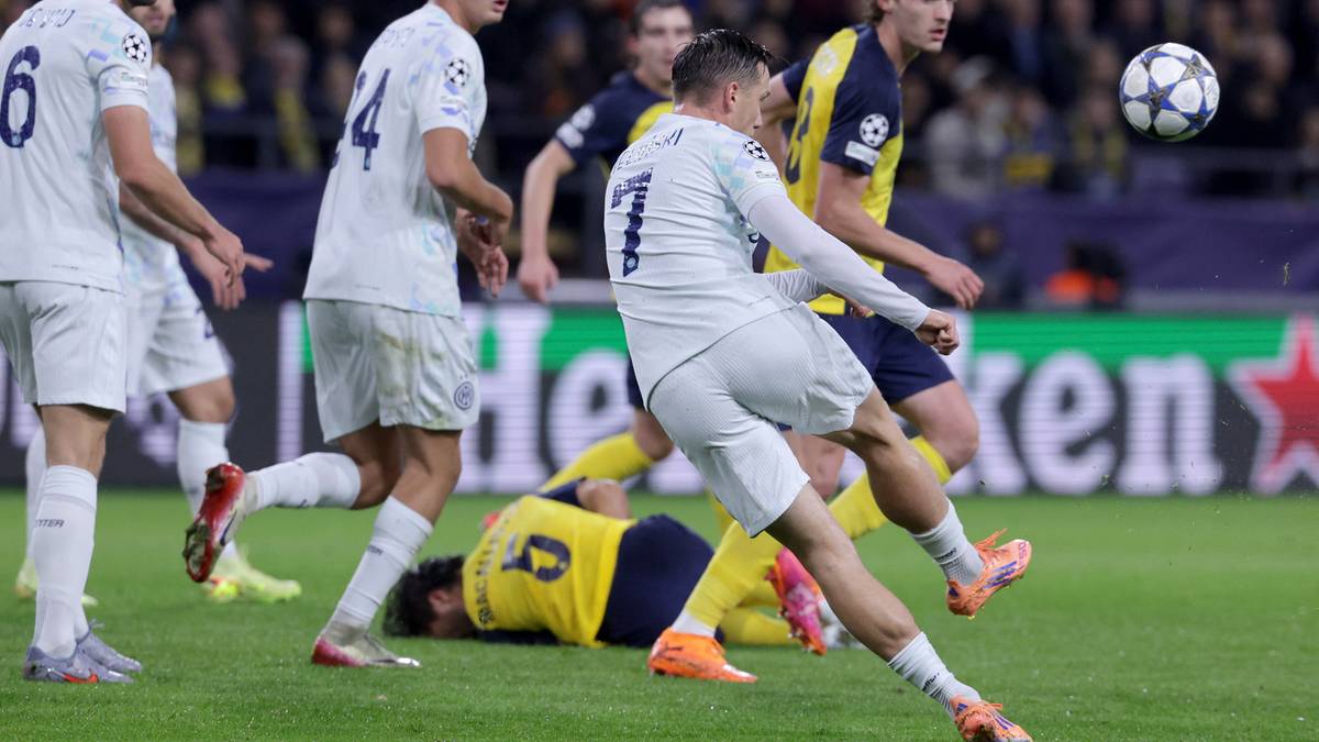 Piłkarz w białej koszulce strzela piłkę do bramki, obok niego leży na ziemi inny zawodnik.