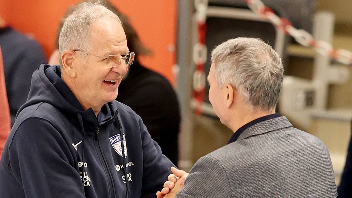 Dwóch mężczyzn w średnim wieku wita się serdecznie, ściskając sobie dłonie. Mężczyzna po lewej ubrany jest w ciemny dres z logo, a mężczyzna po prawej w szarą marynarkę w kratę. Obaj uśmiechają się.