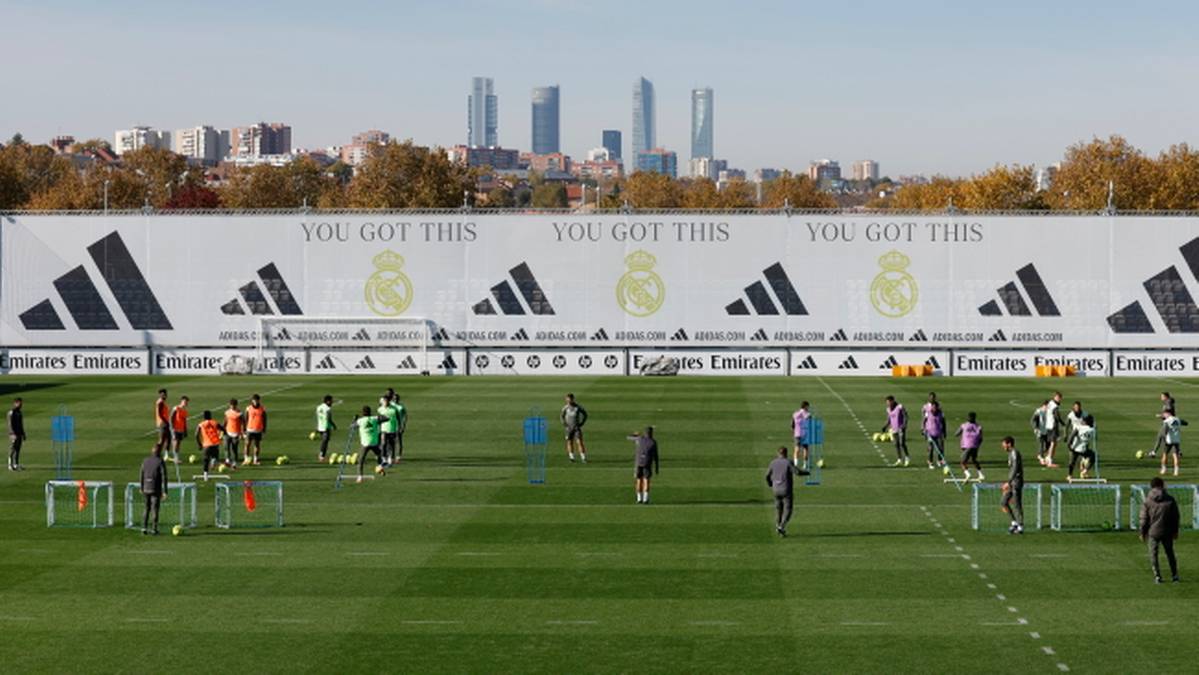 Piłkarze Realu Madryt ćwiczą na boisku treningowym, w tle widoczny baner z logo klubu i panoramą miasta.