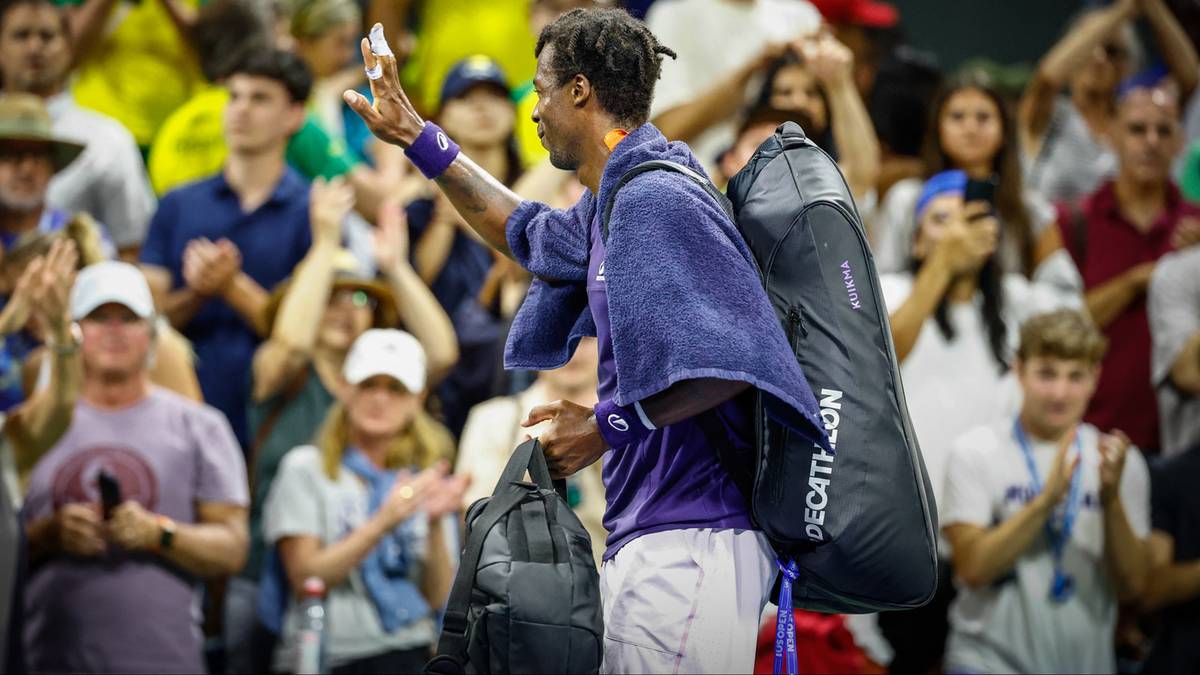 Zamyślony tenisista w stroju sportowym z rakietą na plecach, odwrócony bokiem do widza, stoi na korcie tenisowym przed publicznością.