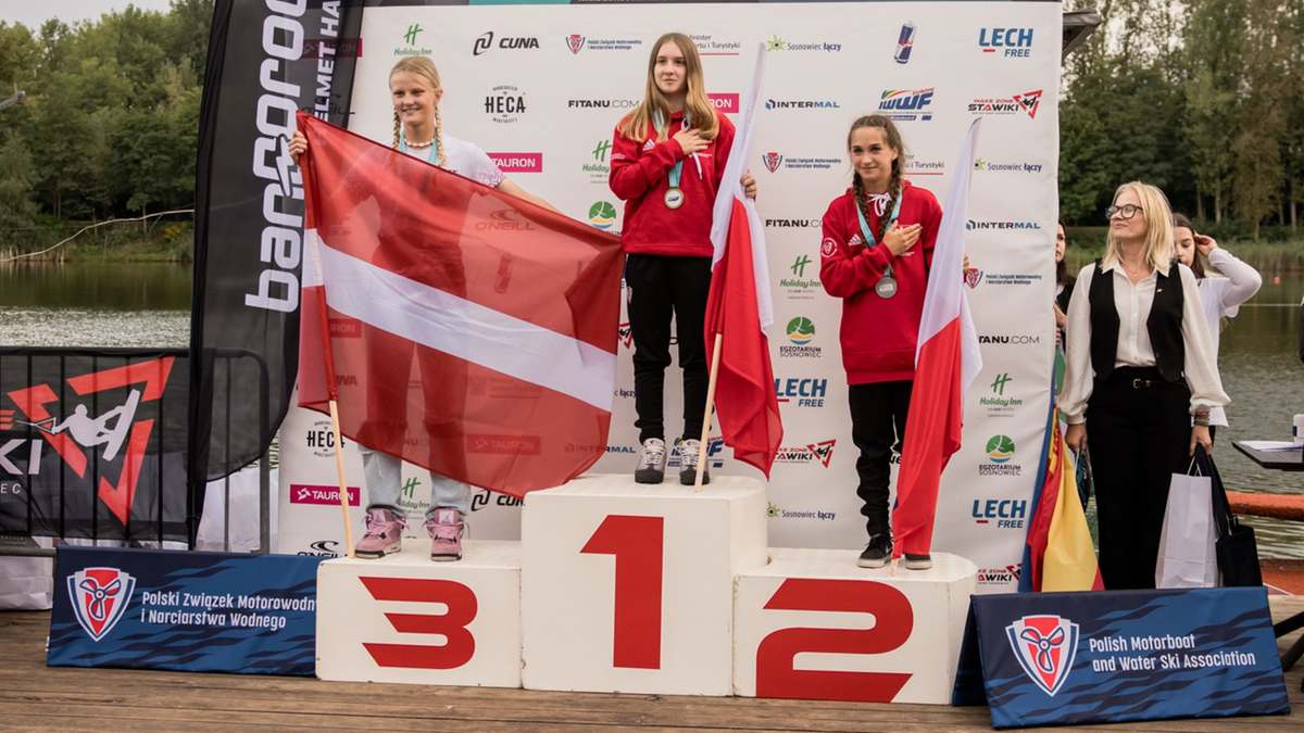 Trzy młode kobiety stoją na podium. Pierwsza z lewej trzyma flagę Łotwy i stoi na miejscu z numerem 3. Druga, w środku, odbiera medal, stojąc na pierwszym miejscu. Trzecia, po prawej, stoi na miejscu z numerem 2 i trzyma flagę Polski. Wszystkie zawodniczki mają na szyjach medale i trzymają dłoń na sercu.
