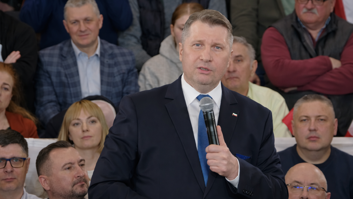 Mężczyzna w garniturze przemawia do mikrofonu, otoczony przez grupę ludzi.