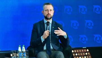 Mężczyzna w garniturze przemawia podczas panelu dyskusyjnego, siedząc na krześle obok kobiety z mikrofonem.