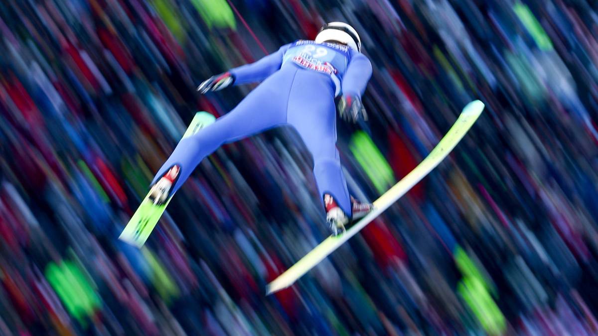 Skoczek narciarski w niebieskim kombinezonie podczas lotu na tle rozmytej publiczności.