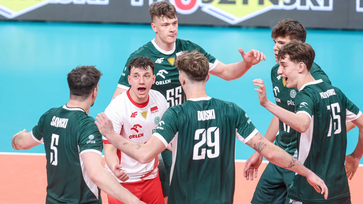 Siedmiu młodych siatkarzy w zielonych strojach sportowych, jeden z nich w białej koszulce z czerwonymi elementami, rozmawia na boisku.