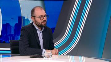 Mężczyzna z brodą i okularami siedzi przy stole, ubrany w marynarkę i koszulę. Na stole znajdują się szklanka z wodą i telefon.