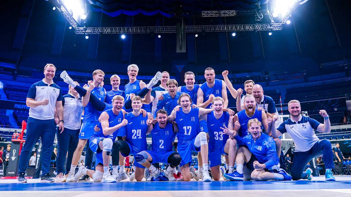 Drużyna siatkarzy w niebieskich strojach pozuje do zdjęcia, trzymając puchar, na tle rozświetlonej hali sportowej. Większość zawodników uśmiecha się, niektórzy podnoszą ręce w geście radości.