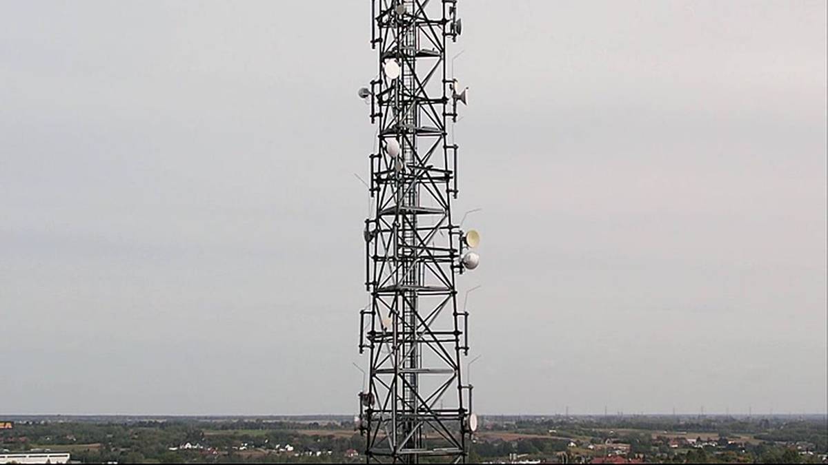 Metalowa wieża telekomunikacyjna z zamontowanymi antenami na tle jasnego nieba i płaskiego krajobrazu.