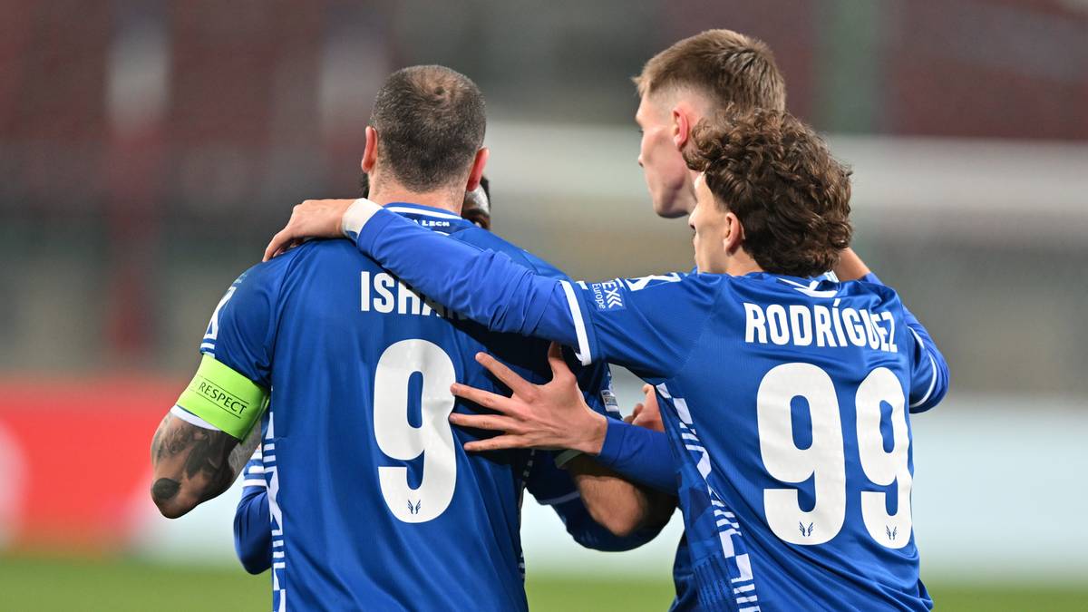 Trzech piłkarzy w niebieskich koszulkach z białymi numerami na plecach obejmuje się po meczu.