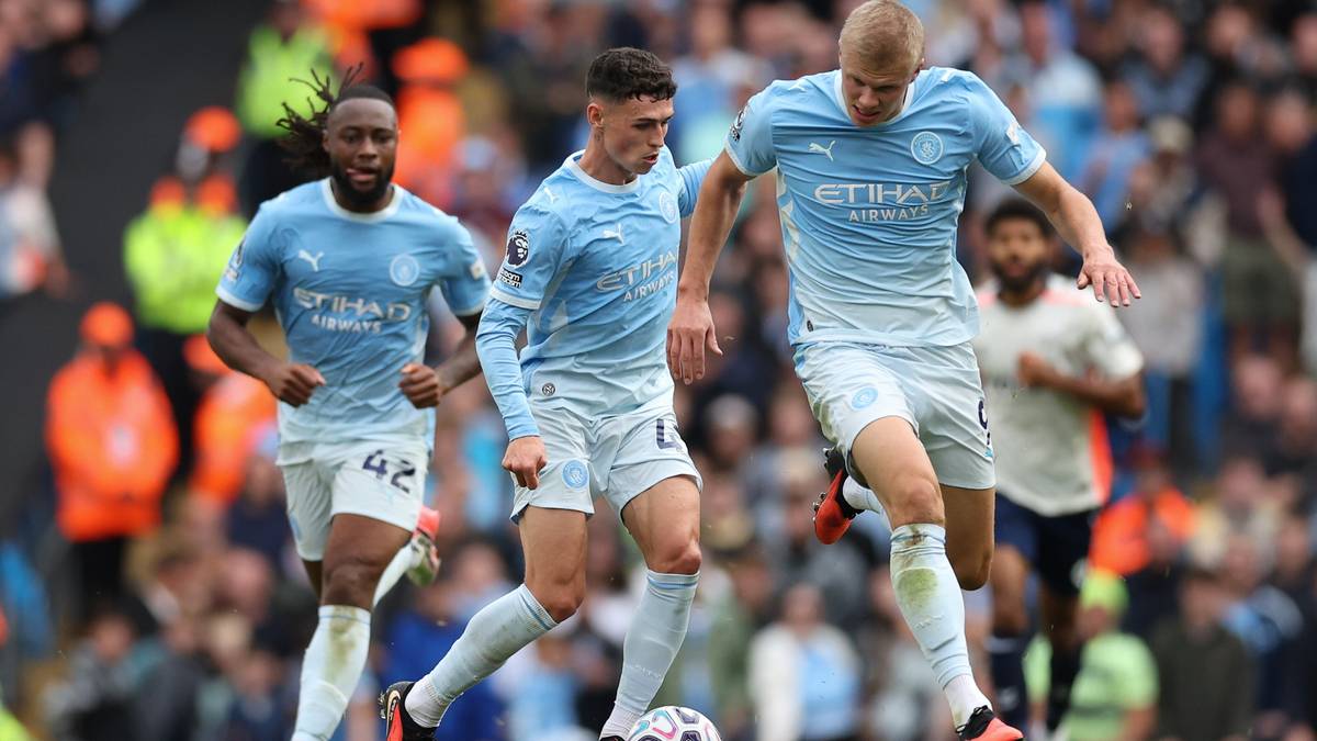 Trzej piłkarze Manchesteru City w jasnoniebieskich strojach biegają po boisku. Jeden z nich drybluje piłkę, podczas gdy dwaj pozostali biegną obok niego. W tle widać kibic&oacute;w.