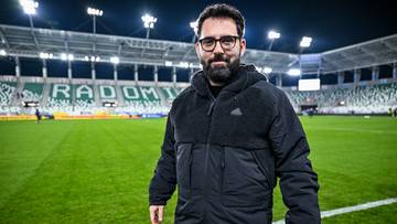 Mężczyzna z brodą i w okularach, ubrany w czarną kurtkę z logo Adidasa, stoi na boisku piłkarskim nocą. W tle widoczne są oświetlone trybuny stadionu z napisem "RADOM".